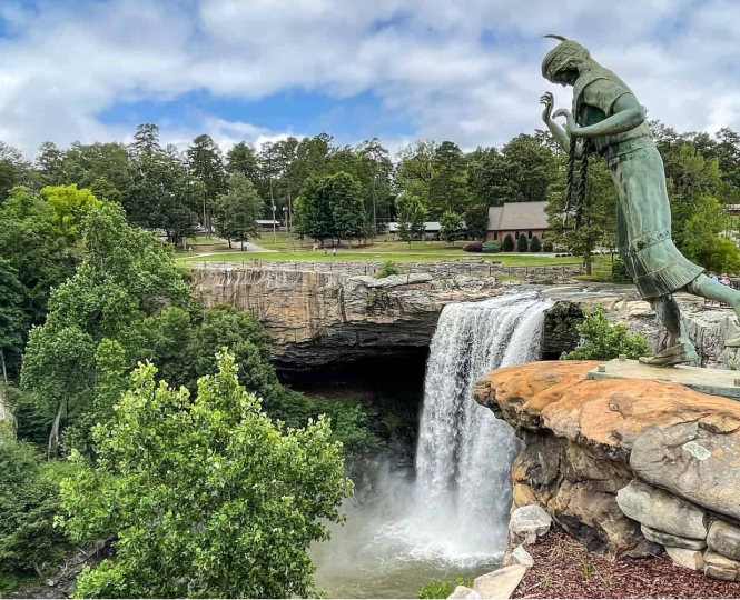 A statue near a waterfall surrounded by trees and a cloudy sky